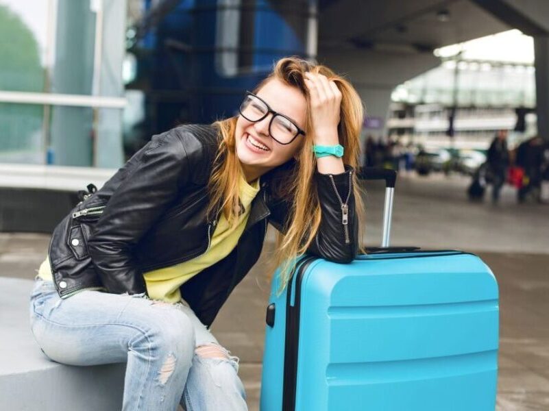 Portrait of pretty girl with long hair in glasses sitting outside in airport. She wears yellow sweater with black jacket and jeans. She leaned to the suitcase and smiling to the camera.